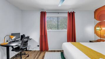 A modern bedroom with a bed, desk, and chair, featuring a window with curtains and a ceiling light.