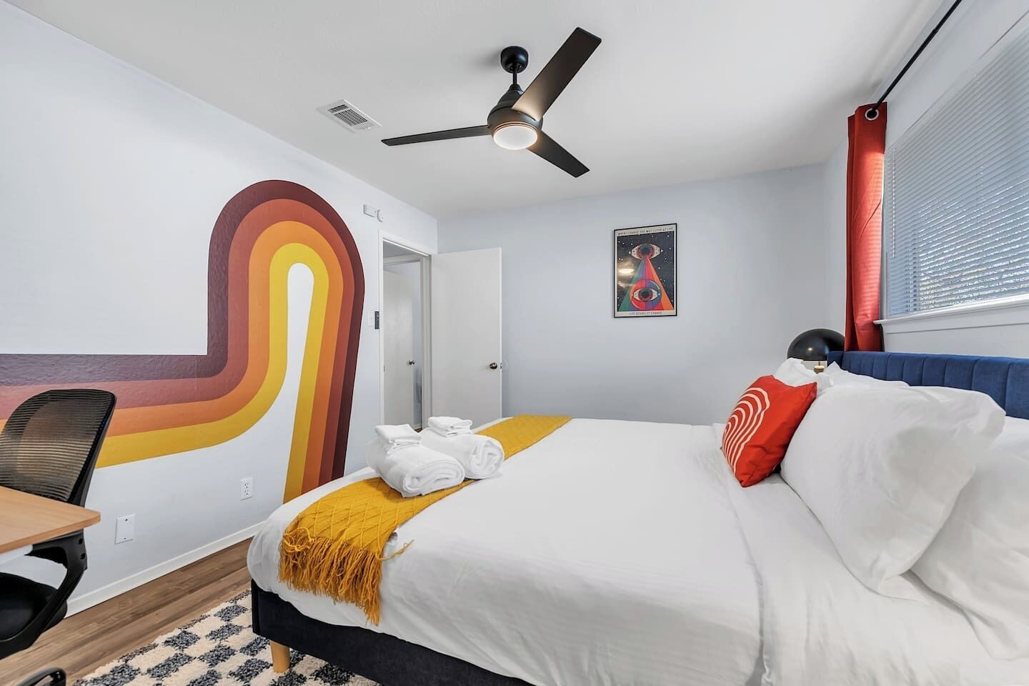 A hotel room with a bed, a desk, a chair, a ceiling fan, and a wall with a colorful abstract design.