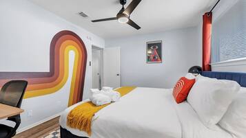 A hotel room with a bed, a desk, a chair, a ceiling fan, and a wall with a colorful abstract design.