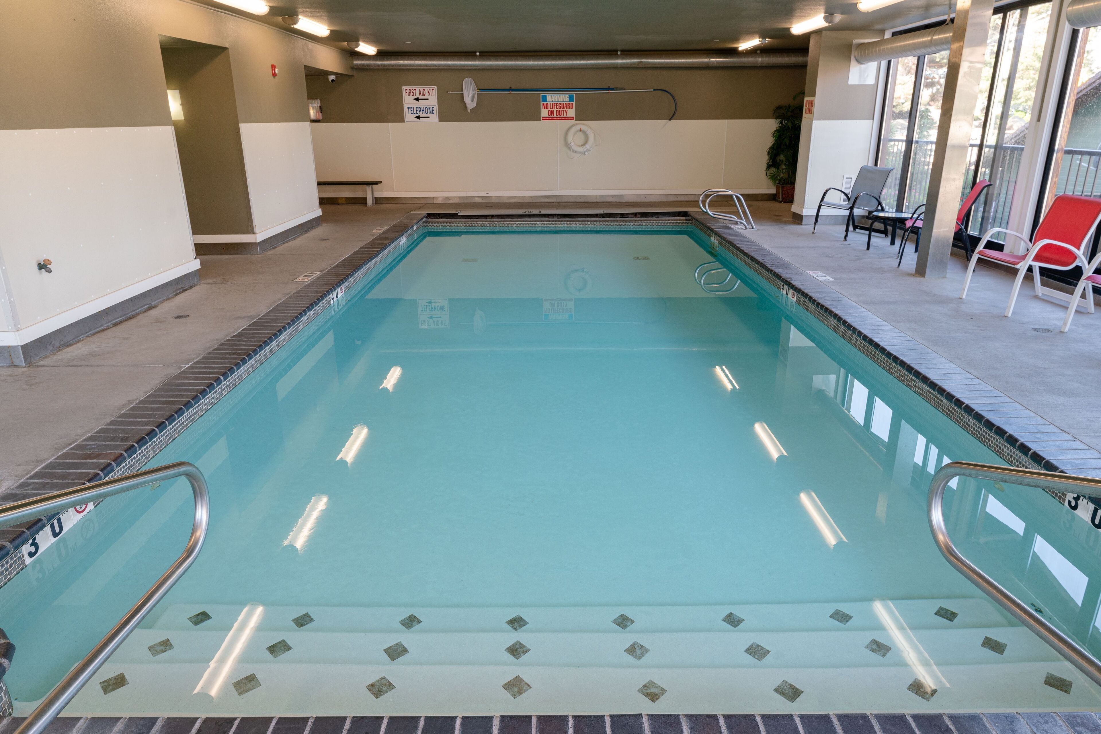 En indendørs swimmingpool med klart vand, en redningsbadsstol og et skilt der viser placeringen af et førstehjælpsrum.