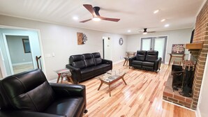 A spacious living room with two black leather sofas, a glass coffee table, and a fireplace with a brick surround. - 3 Bedroom House at Pine Forest Hideaway Near UNC (Chapel Hill)