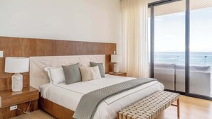 A modern bedroom with a large bed, bedside lamps, a bench, and a view of the ocean through a sliding door.