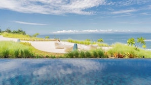 A pool with a view of the ocean and a sandy area with grass and palm trees.