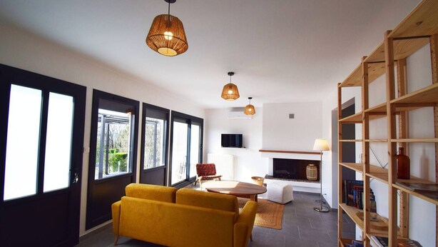 A modern living room with a sofa, a coffee table, and a fireplace. There are large windows and a bookshelf. - Gîte moderne avec clim Bet Ceou - proche Agen (COLAYRAC ST CIRQ)