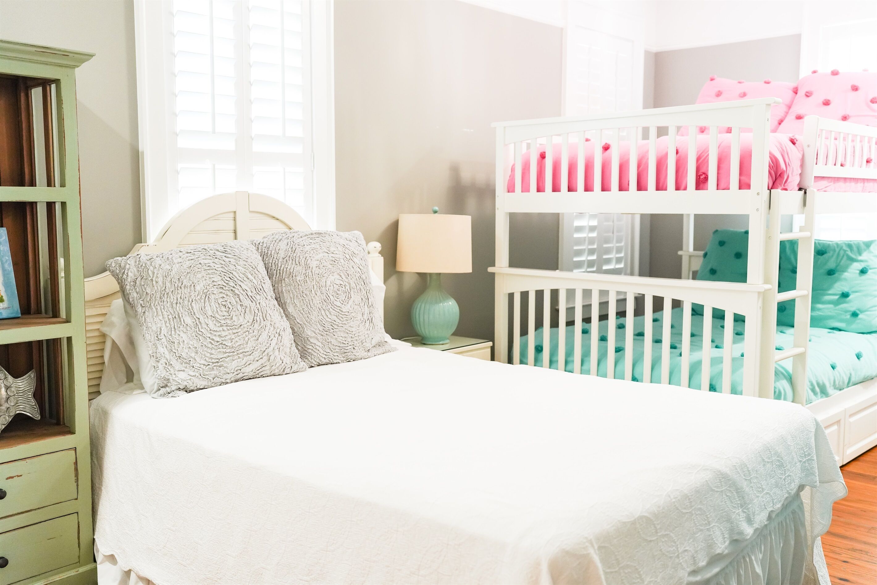 A bedroom with a white bed, a green nightstand, a white crib, and a window with blinds.