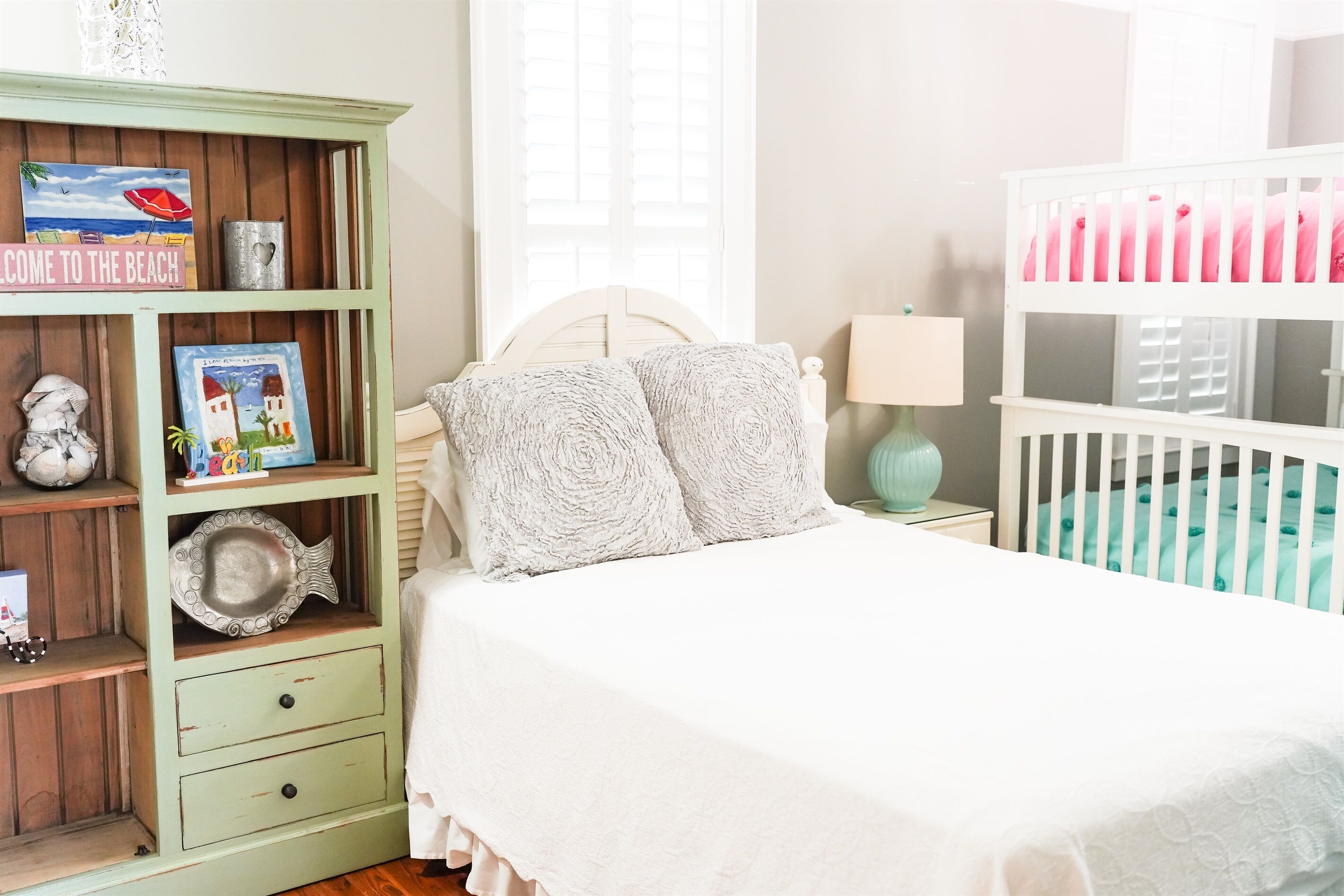A bedroom with a crib, a green dresser with decorative items, a white bed with a white bedspread, and a lamp on a bedside table.