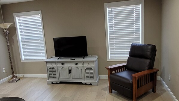 A living room with a television on a white cabinet, a leather chair, and two windows with blinds.