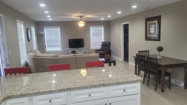 A modern kitchen with granite countertops, a dining area with a table and chairs, a living room with a sofa and a TV, and a ceiling fan.