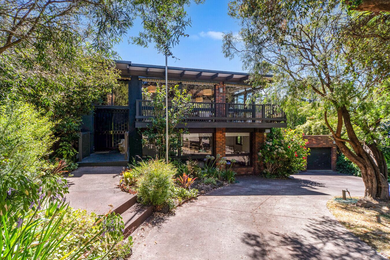 A two-story house with a balcony, surrounded by lush greenery and a paved driveway.