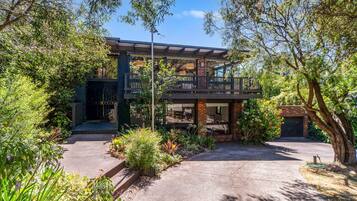 A two-story house with a balcony, surrounded by lush greenery and a paved driveway.