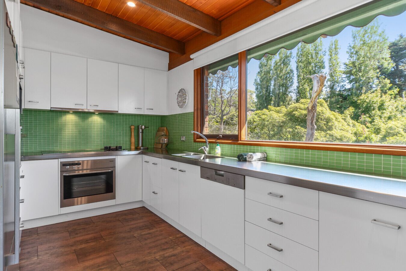 A modern kitchen with white cabinets, a green tiled backsplash, a stainless steel oven, and a window with a view of trees.