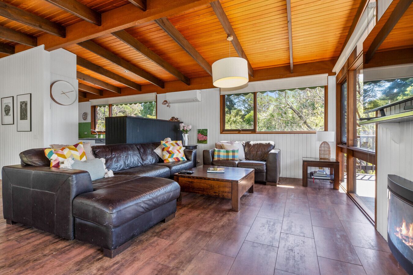 A spacious living room with a fireplace, leather sofas, wooden flooring, and large windows offering a view of the outdoors.