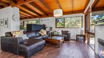 A spacious living room with a fireplace, leather sofas, wooden flooring, and large windows offering a view of the outdoors.