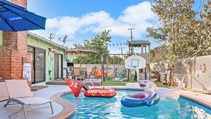 A backyard with a swimming pool, inflatable pool toys, and a basketball hoop. - Pirates Cove: Heated Pool, Arcades, Playground and more! (Garden Grove)