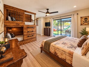 A bedroom with a wooden bunk bed, a large bed with a nautical-themed duvet, a ceiling fan, and a view of a slide and outdoor area.