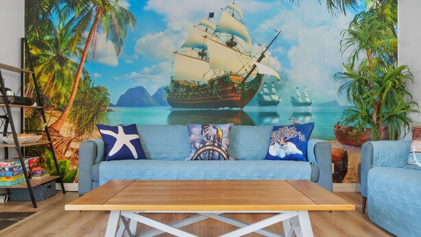 A room with a large tropical mural, a blue sofa, and a wooden coffee table. - Pirates Cove: Heated Pool, Arcades, Playground and more! (Garden Grove)