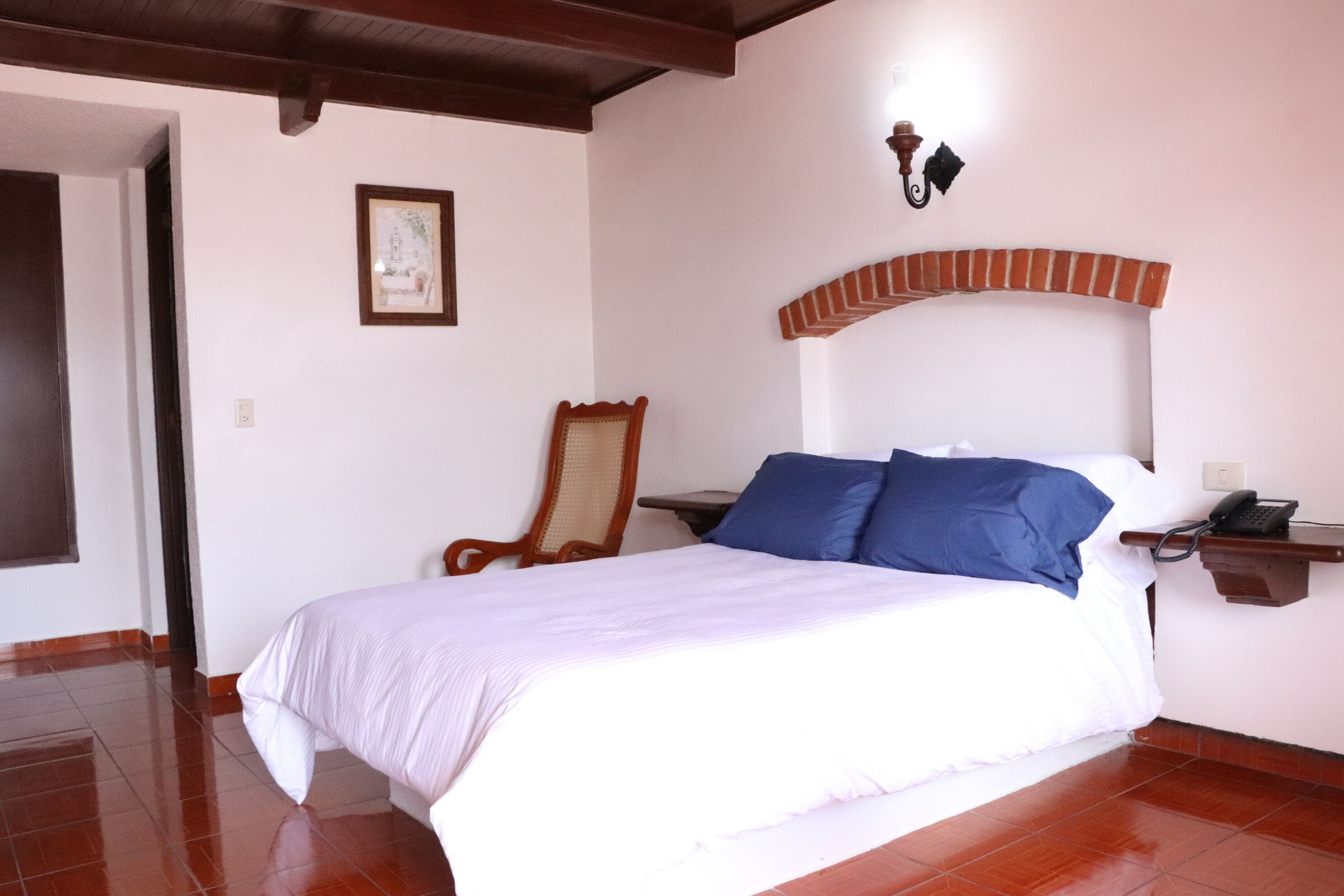 Un dormitorio con una cama, una silla, una mesita de noche con un teléfono, y una lámpara de pared.