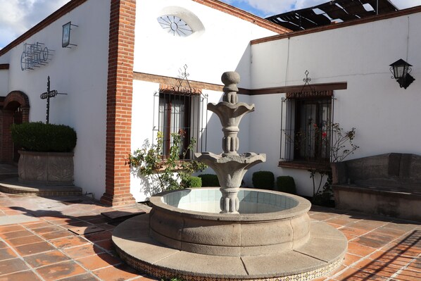 A courtyard with a central fountain, a white building with a clock, and a tiled floor. - Hotel Cuamanco (Huamantla)