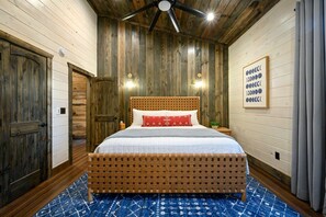 Ein Schlafzimmer mit hölzernem Kopfteil, einem Bett mit weißer Bettwäsche, einem roten Kopfkissen und einem Deckenventilator. An der Wand hängt ein gerahmtes Bild.
