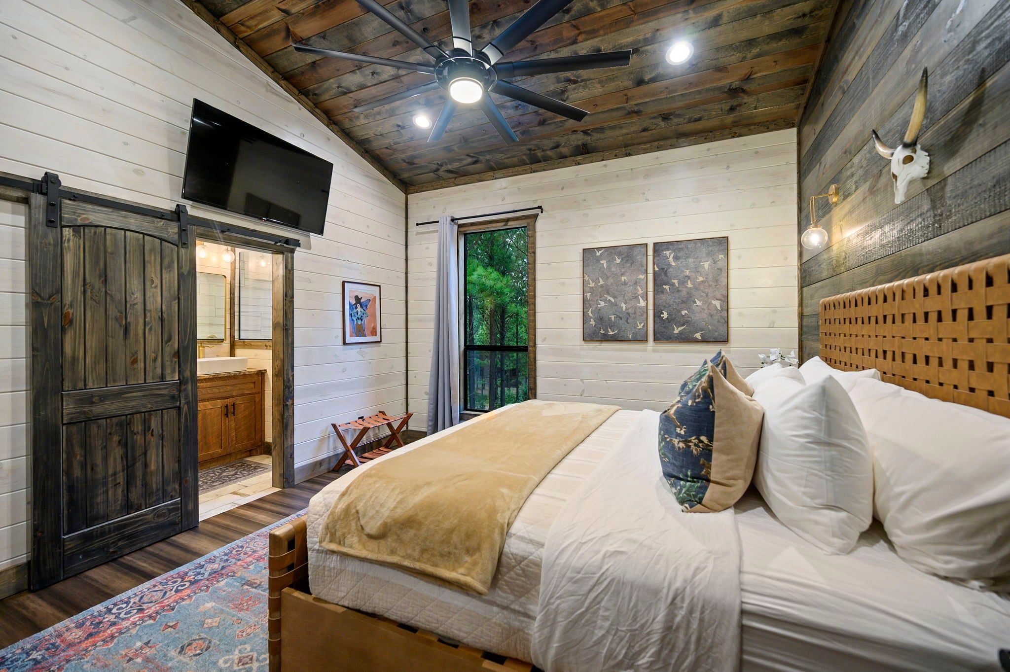 A bedroom with a bed, a ceiling fan, a mounted TV, and a large window.