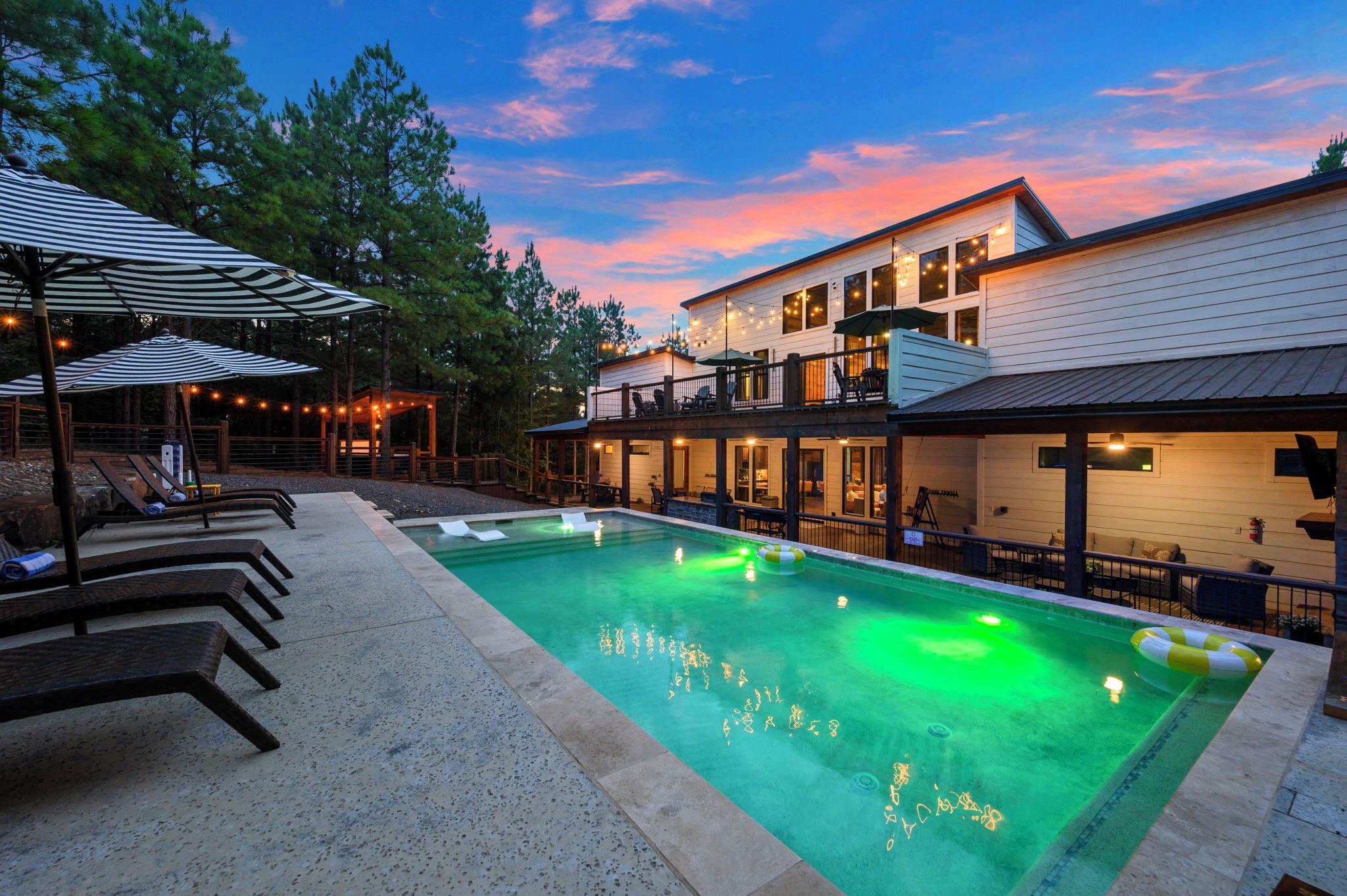 A swimming pool with lounge chairs and umbrellas, a covered seating area, and a building with outdoor lighting.