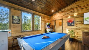 Ein Billardtisch in einer Holzhütte mit Holzwänden und Fenstern, die auf einen Wald blicken.