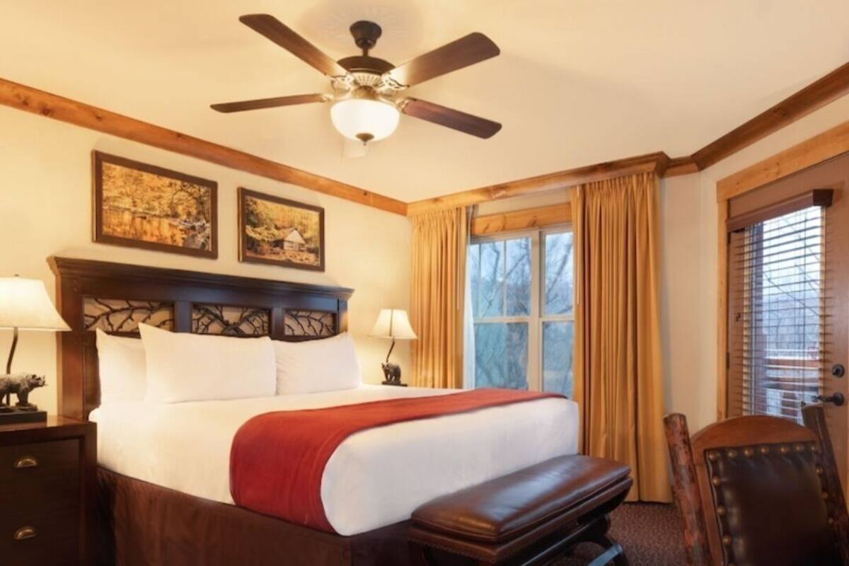 A large bed with a wooden headboard, two bedside lamps, a red throw blanket, a wooden bench, a leather chair, and a ceiling fan.