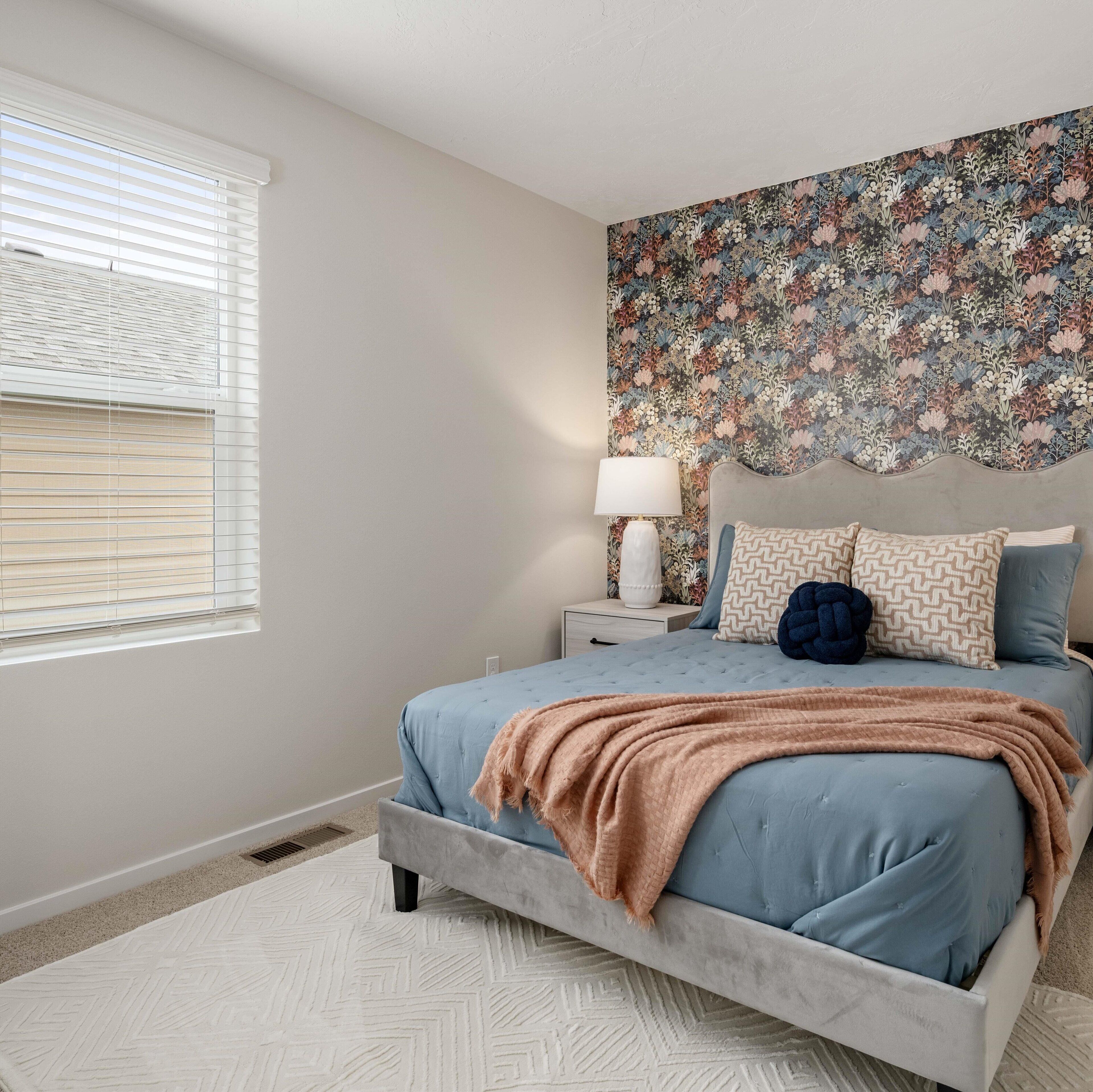 A bedroom with a bed, a nightstand, a lamp, a floral wallpaper, and a window with blinds.
