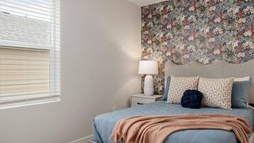 A bedroom with a bed, a nightstand, a lamp, a floral wallpaper, and a window with blinds.