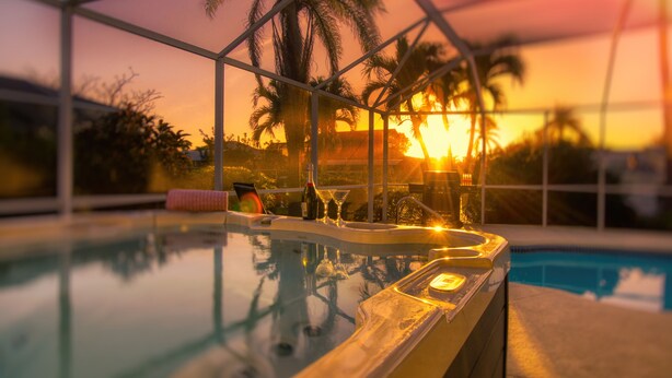 A pool area with a sunset view, palm trees, and a bar with glasses and bottles. - VILLA RIVERBREEZE – HEATED POOL & SPA | GULF ACCESS | BOAT RENTAL + TOURS VOUCHER INCL. (Cape Coral)