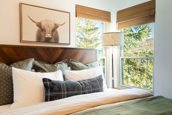 Une chambre à coucher avec un lit, des oreillers, un tableau encadré, une fenêtre donnant sur un beau paysage et une lampe.