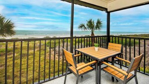 Un balcón con una mesa y sillas de madera, con vista a una playa con palmeras y cielo despejado.