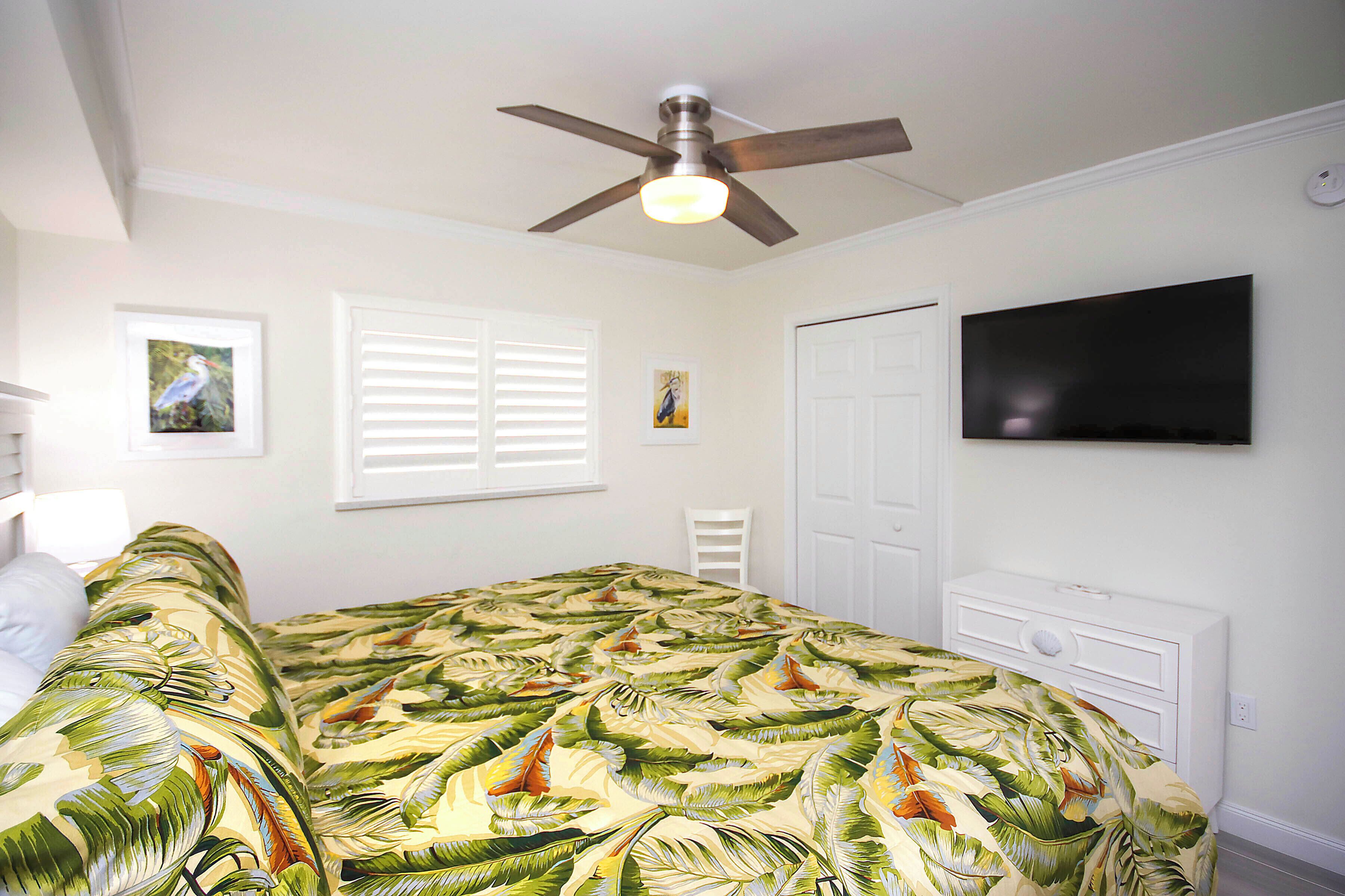 A bedroom with a bed, a ceiling fan, a TV, and a window with blinds.