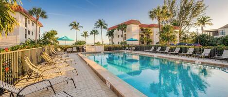 A swimming pool with lounge chairs and umbrellas, surrounded by palm trees and a building with a red roof.