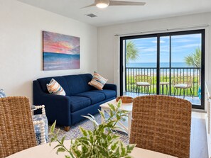 Un salon avec un canapé bleu, des fauteuils en osier, un tableau et un balcon offrant une vue sur l’océan.