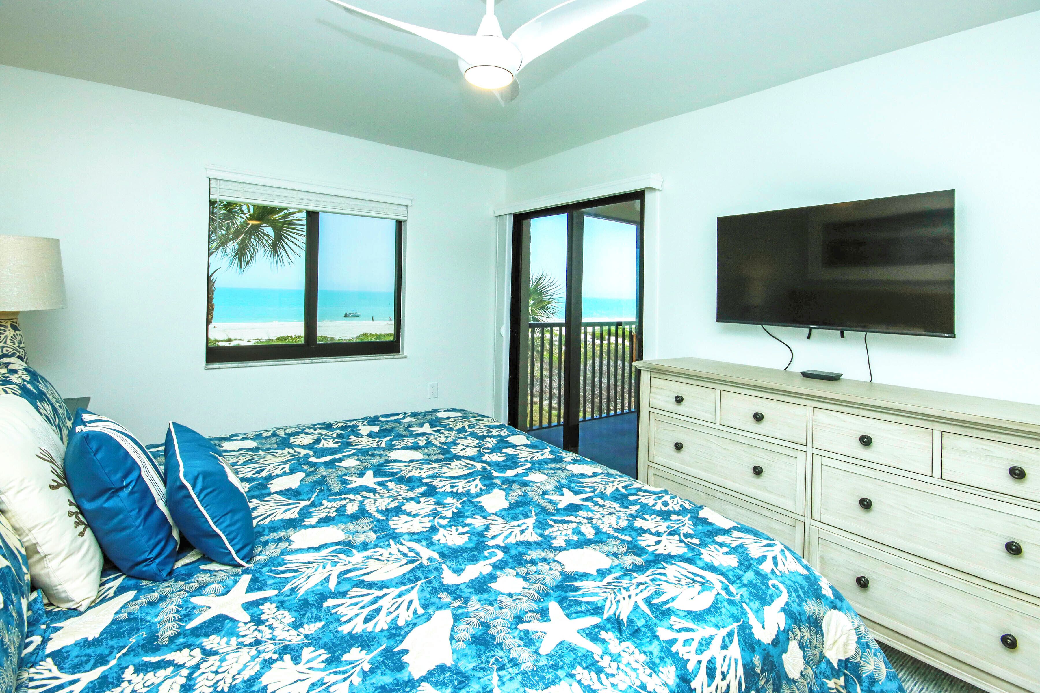Een slaapkamer met een bed, een dressoir, een televisie en een raam met uitzicht op het strand.