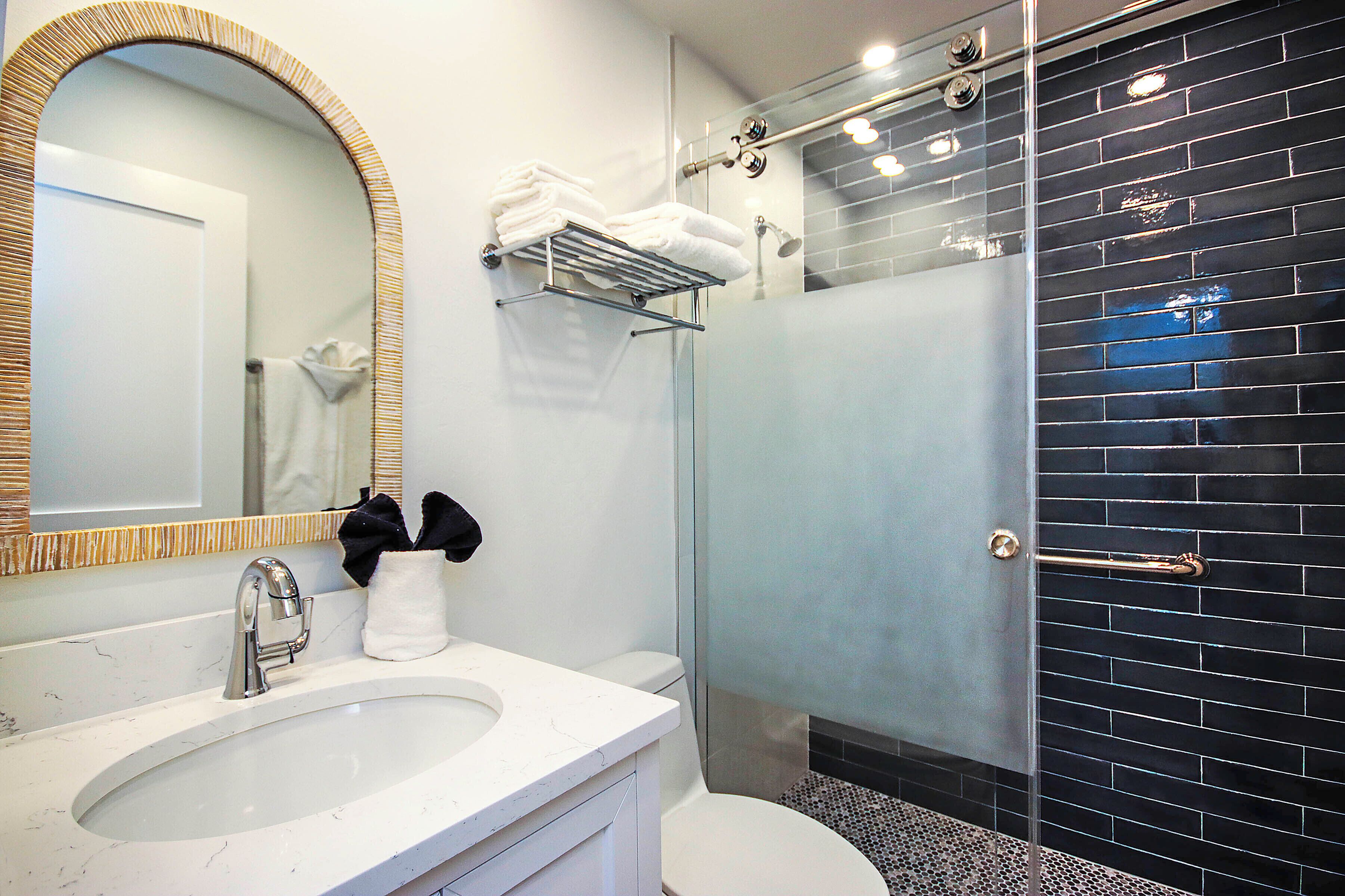 Une salle de bain moderne avec un lavabo blanc, un grand miroir, une douche en verre et un mur recouvert de carreaux noirs.