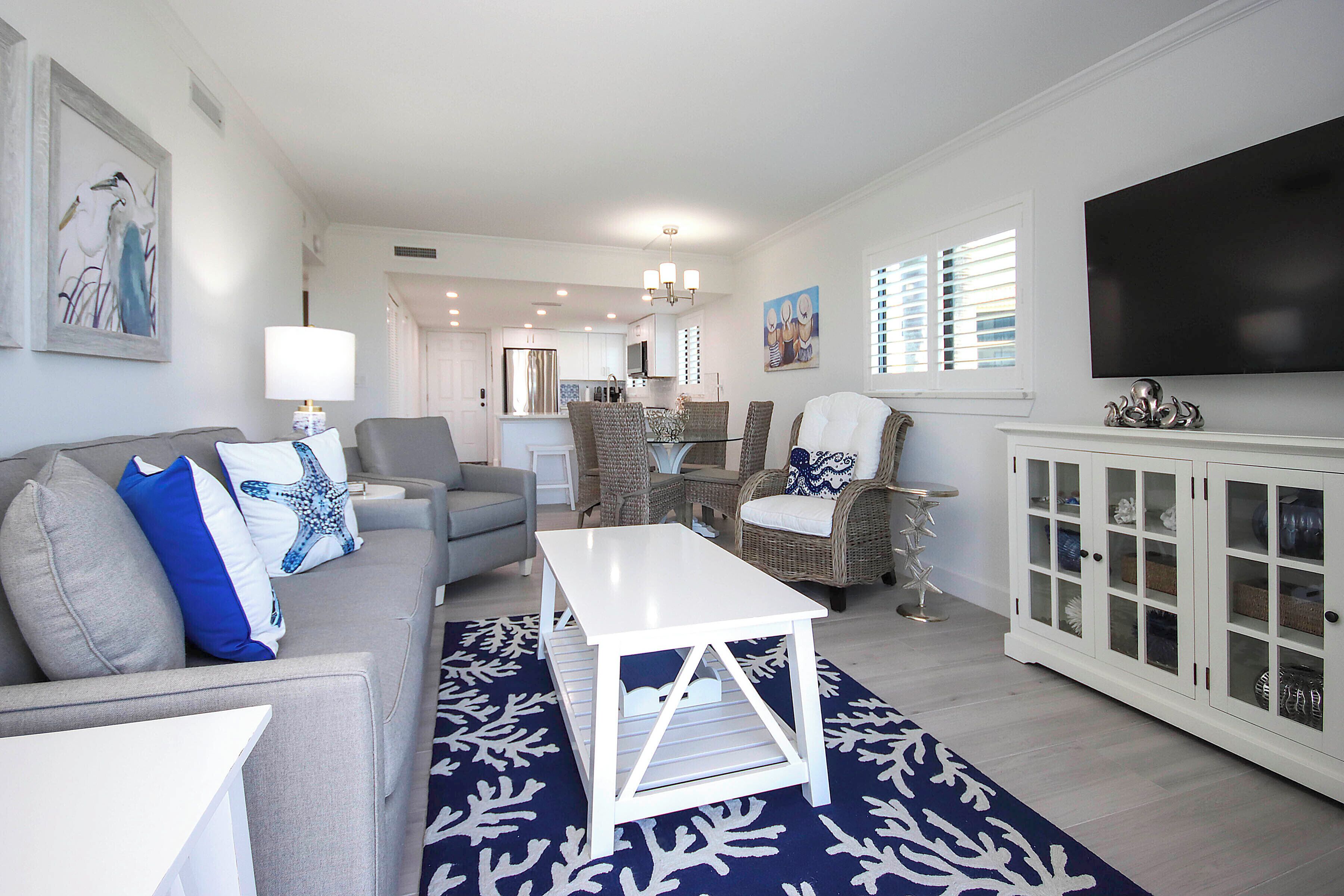 A living room with a grey sofa, white coffee table, wicker chairs, a white cabinet with glass doors, and a flat-screen TV mounted above the cabinet.