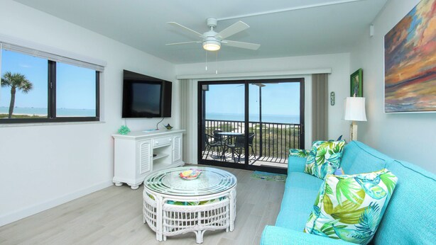 Habitación con balcón, ventilador de techo, televisor, mesa redonda y sofá azul con almohadones tropicales.