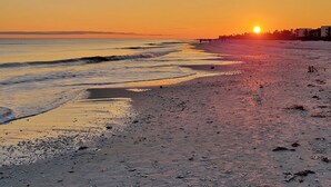 A beach at sunset with the sun low on the horizon, waves gently lapping the shore, and a distant shoreline with buildings.