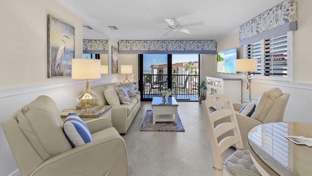 A spacious living room with a sofa, chairs, a coffee table, and a TV. There is a balcony with a view of the ocean.