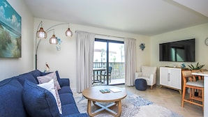 Ein Wohnzimmer mit einer Couch, einem runden Couchtisch, einem Fernseher und einem Balkon mit Vorhängen.