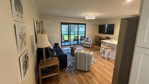 Un vaste espace de vie comprenant un canapé, un pouf et une table à manger avec des chaises. Il y a également un coin cuisine équipé d’un réfrigérateur et d’un téléviseur à écran plat fixé au mur.