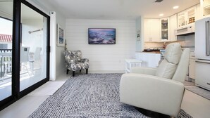 A modern living room with a large window, a ceiling fan, a patterned rug, a white sofa, and a kitchen area with white cabinets and appliances. - 1BR ocean view condo on Sanibel Island Steps from the beach - Sandalfoot 3B3 (Sanibel Island)