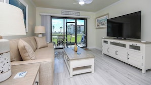 Una sala de estar con un sofá color beige, una mesa de centro, un televisor y un ventilador de techo. Hay un letrero que dice "Welcome to the Beach" y se ve una vista de palmeras y edificios a través de las puertas de vidrio.
