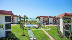 A pool area with a shuffleboard court, surrounded by buildings with red-tiled roofs and a clear view of the ocean. - 3rd Story Sanibel Condo With Coastline Views - Sandalfoot 3C3 (Sanibel Island)