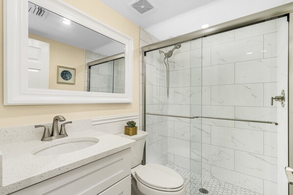 Une salle de bain avec un lavabo blanc, un miroir, des toilettes et une douche séparée par une paroi de verre.