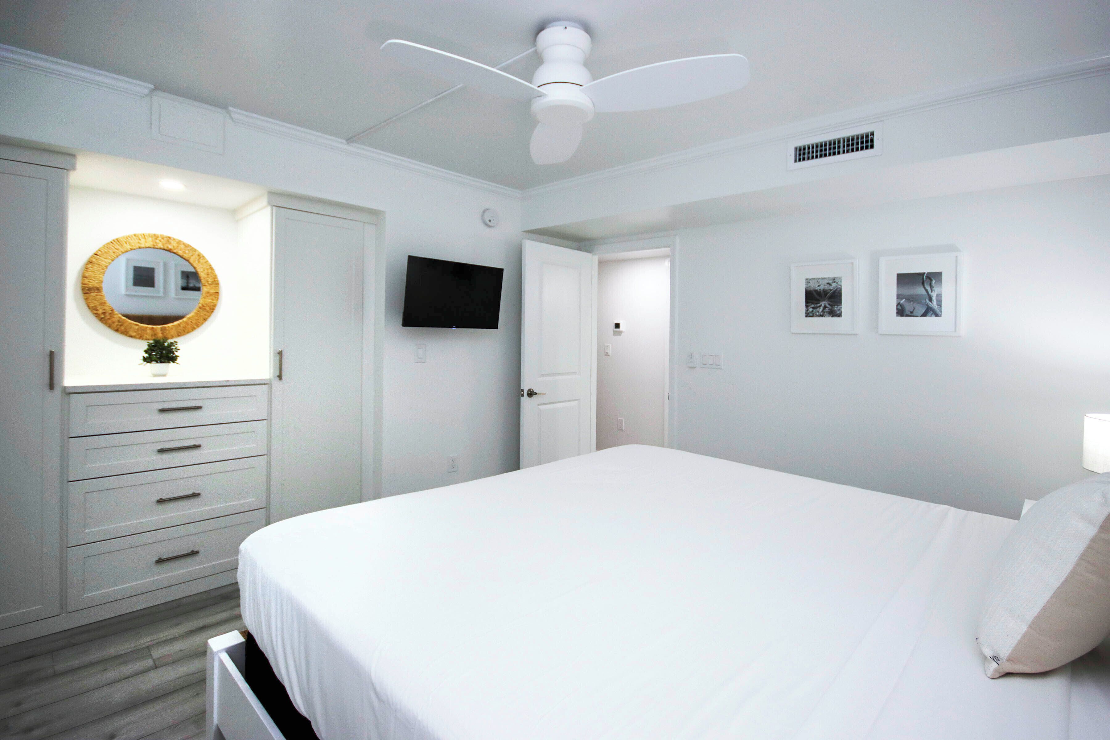 Un dormitorio con cama, ventilador de techo, televisor, espejo y cómoda.