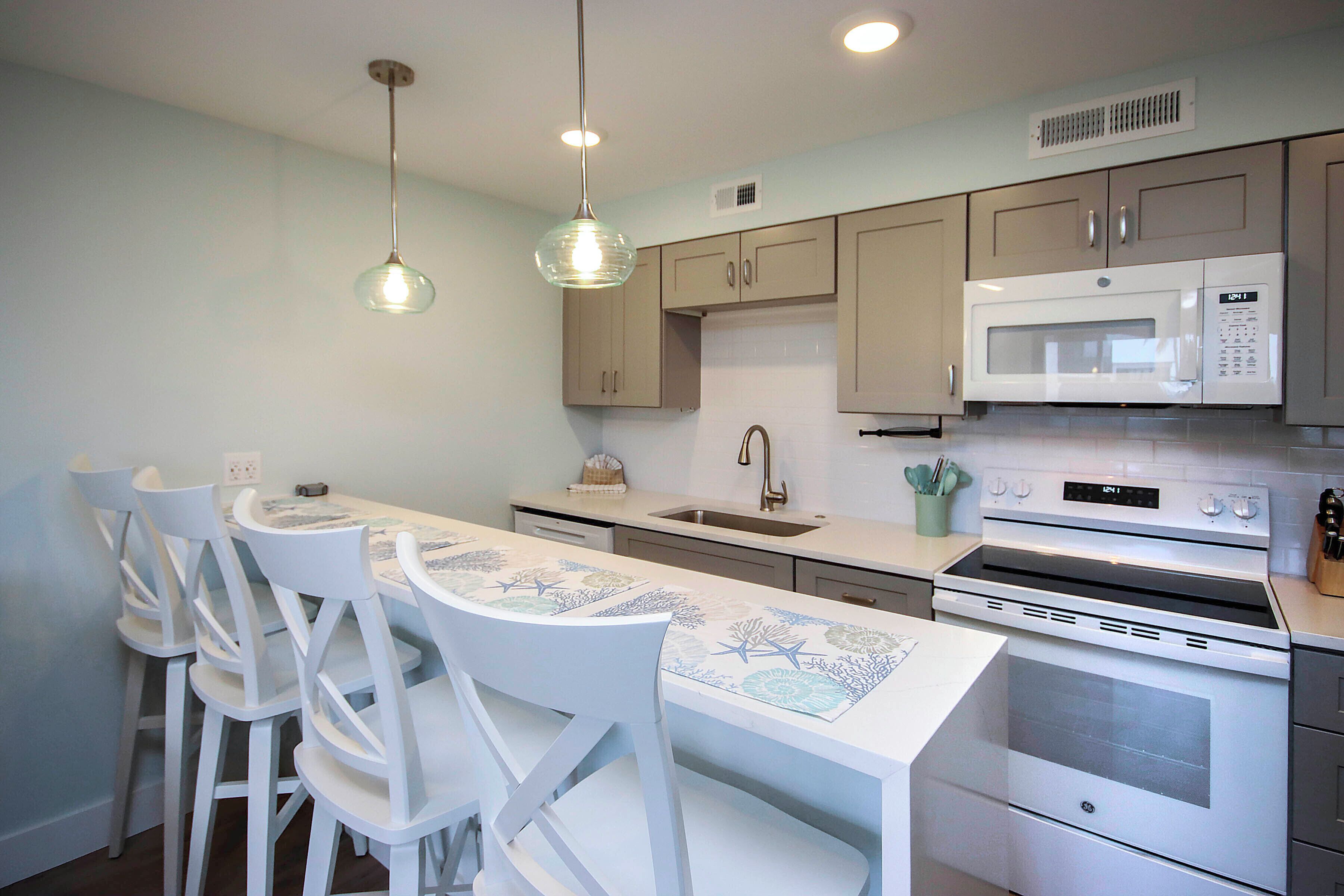 A kitchen with white cabinets, a white countertop, a white dining table with chairs, a microwave, an oven, and pendant lights.