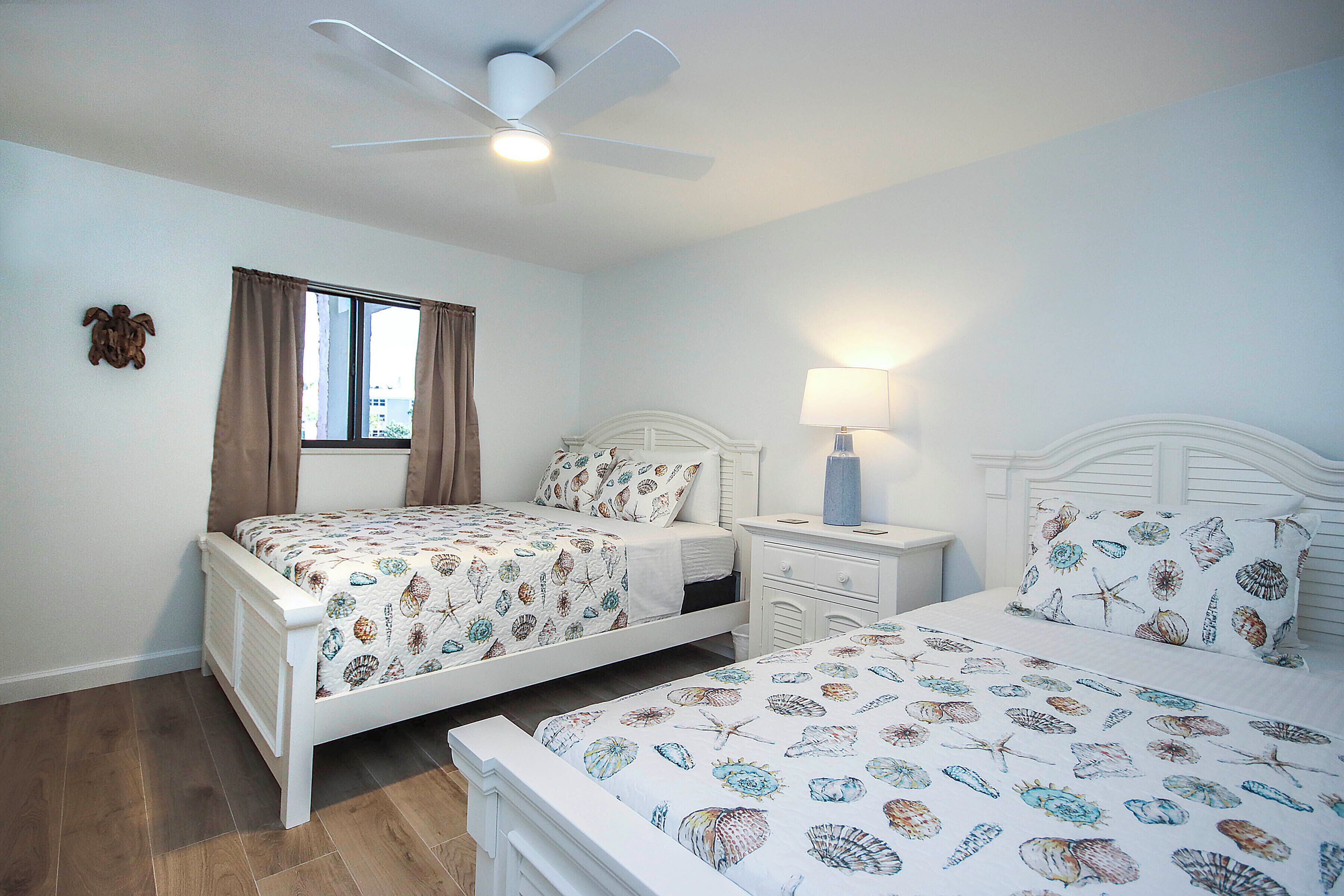 A bedroom with two beds, a ceiling fan, a window with curtains, and a bedside lamp.